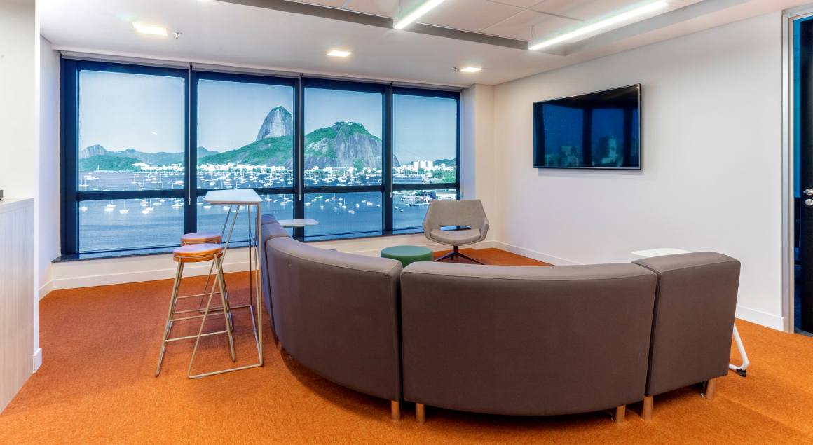 Living area in the office with a large window overlooking Pão de Açúcar. In the foreground, a curved sofa and high stools are arranged around a counter, creating a cozy and sophisticated environment.