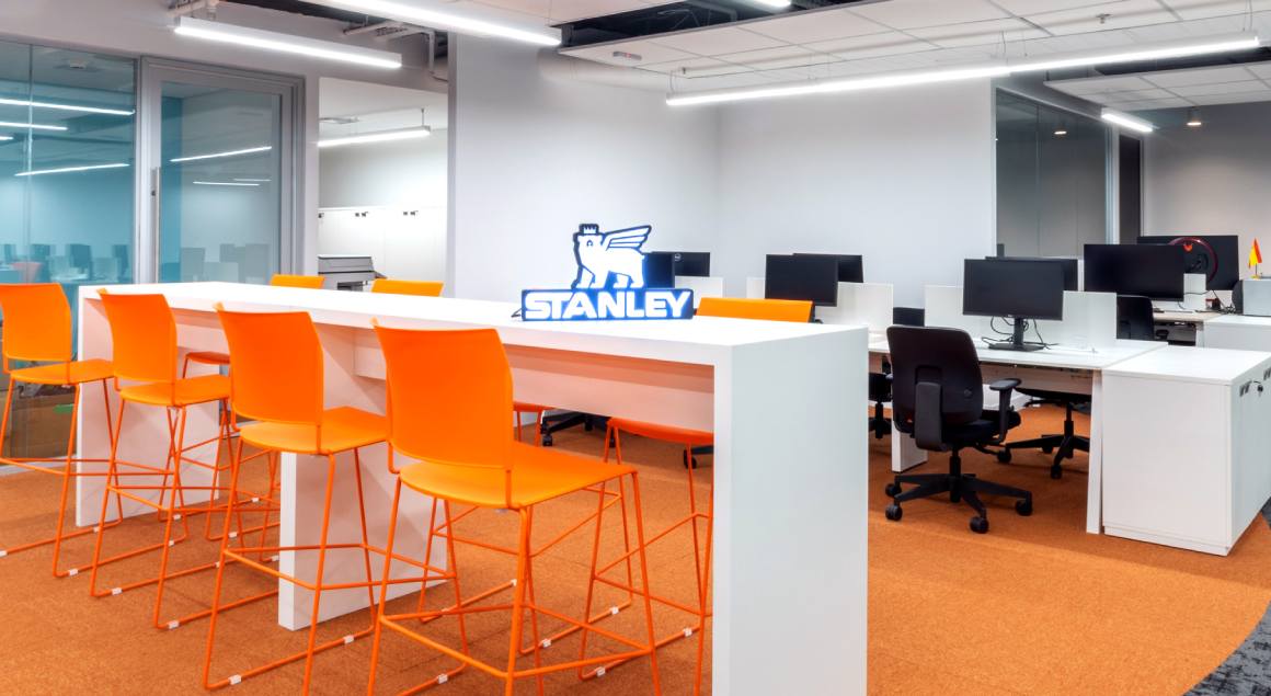 In the background, a modern office with tables and chairs arranged in workstations, a high bench with tall orange chairs, creating a communal area. The Stanley brand is visible, reinforcing the identity of the space.