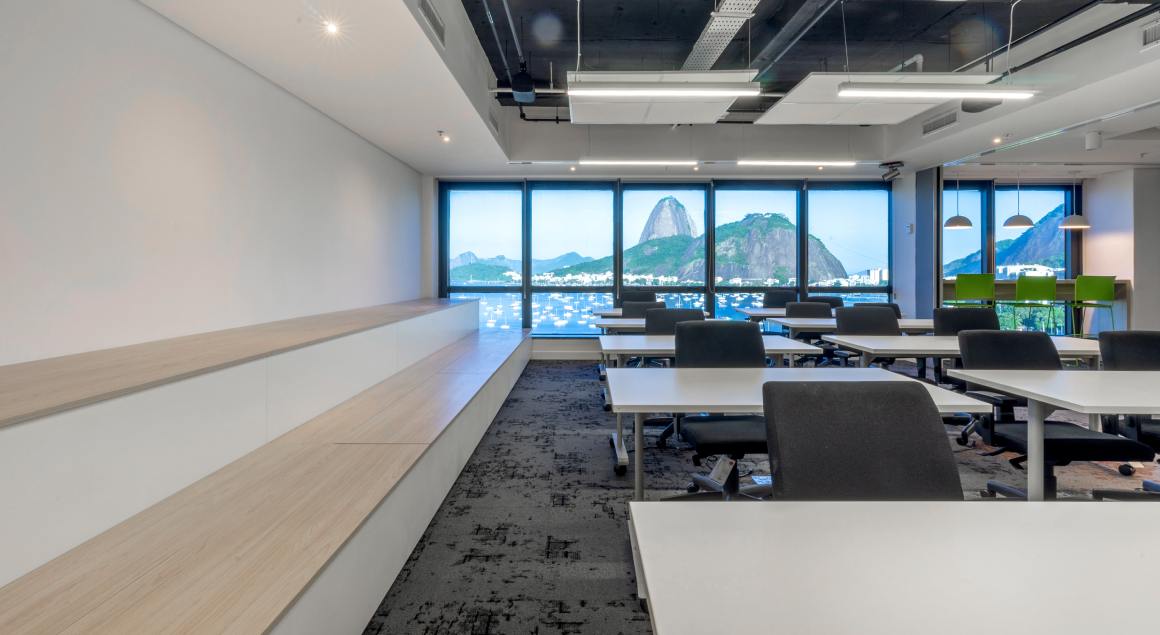 Várias mesas de escritório em um ambiente iluminado por luz natural, com amplas janelas que oferecem vista do Pão de Açúcar no Rio de Janeiro ao fundo.